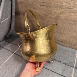 May include: A hammered brass bucket with a curved handle and three claw feet. The bucket has a flared rim and a hinged handle attachment. The brass has a warm, golden hue, and the bucket appears to be vintage.