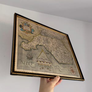 May include: Framed antique map with a black and gold frame. The map features detailed illustrations of land and sea, with text in Latin. A compass rose is visible in the lower right corner. The map is held by a person.