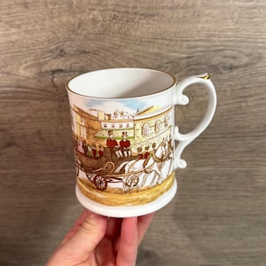 Puede incluir: Taza de cerámica blanca con una ilustración detallada de un carruaje tirado por caballos y figuras con ropa de época. La taza tiene un asa y un borde dorados, y el diseño envuelve la taza.