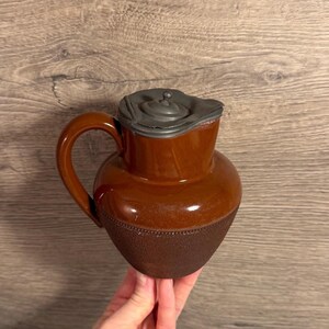 Op de afbeelding: Een bruine keramische kan met een handvat en een tinnen deksel. De kan heeft een bolvormige body en een getextureerd onderste gedeelte. Het handvat is gebogen en het deksel heeft een kleine knop. De kan is waarschijnlijk voor het serveren van dranken.