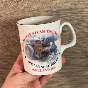 May include: White ceramic mug with a black rim and handle. The mug features a red and black image of a steam engine with the text "Ross-on-Wye Steam Engine Society 40th Annual Rally Welland 2004".