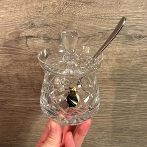 Puede incluir: Un azucarero de vidrio transparente con tapa y una cucharita. El tazón tiene un diseño de vidrio cortado.