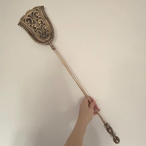 Op de afbeelding: Een gouden haardgereedschap met een sierlijk, decoratief hoofd en een lange, slanke steel.