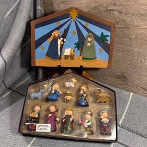 Könnte beinhalten: Ein Weihnachtskrippenset mit bemalter Kulisse und Figuren. Die Kulisse zeigt einen Stern, Figuren und Palmen. Das Set enthält Figuren von Maria, Josef, Jesus und anderen Figuren. Das Set befindet sich in einer blauen Box.
