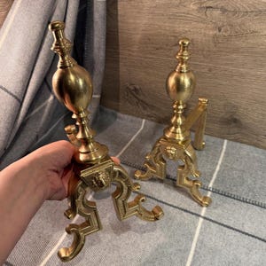 Puede incluir: Par de morillos de chimenea antiguos de latón. Cada morillo presenta una base ornamentada y un soporte central con un diseño bulboso. Los morillos son de color dorado cálido y se exhiben sobre una manta a cuadros gris y blanco.
