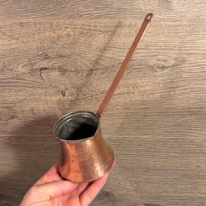 May include: A copper coffee pot with a long handle and a hammered texture. The pot has a rounded base and a narrow spout. The handle has a small hole at the end for hanging. The pot is held in a hand.