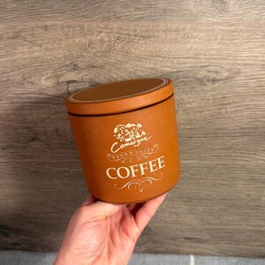 May include: A cylindrical terracotta coffee canister with a matching lid. The front features a white graphic of a horse above the text Camargue Terre Cuite and the word COFFEE in large capital letters with decorative flourishes below.