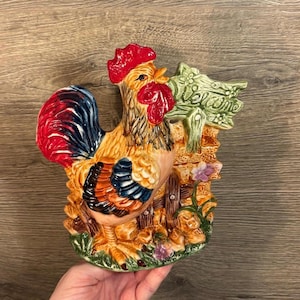 Könnte beinhalten: Eine Keramik-Hahnenfigur mit einem rot, blau und gelb bemalten Design. Der Hahn steht neben einem Holzzaun mit einem grünen Schild, auf dem "Willkommen" steht.