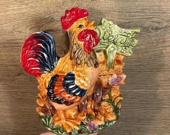 Peut inclure: Une figurine de coq en céramique avec un motif peint rouge, bleu et jaune. Le coq se tient à côté d'une clôture en bois avec une pancarte verte qui dit "Bienvenue".