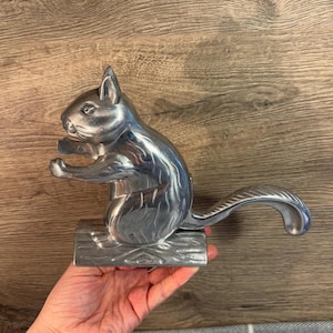 May include: A silver squirrel nutcracker with a detailed design. The squirrel is sitting on a log base and has a long tail that acts as the nutcracker.