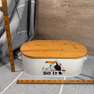 May include: An oval white metal storage container with a bamboo lid and two side handles. The front features a toucan graphic with the text "toucan DO IT". A wooden folding ruler shows the container is approximately 13 inches wide and 6 inches tall.