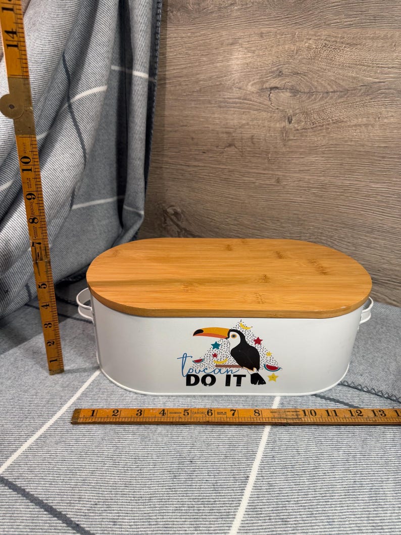 May include: An oval white metal storage container with a bamboo lid and two side handles. The front features a toucan graphic with the text "toucan DO IT". A wooden folding ruler shows the container is approximately 13 inches wide and 6 inches tall.