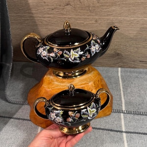May include: A black ceramic teapot and matching sugar bowl set featuring a gold-trimmed lid, handle, and base. The pieces are decorated with a botanical pattern of white flowers, green leaves, and small red berries painted on the glossy black surface.