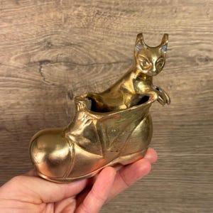 May include: A gold-colored metal figurine of a cat sitting inside a shoe. The shoe is a boot with a pointed toe and a high heel.