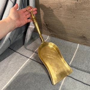 Könnte beinhalten: Eine Vintage-Schaufel aus Messing mit langem Griff und Ring oben. Die Schaufel hat eine gebogene Form und eine polierte Oberfläche. Der Griff hat dekorative Details. Die Schaufel wird wahrscheinlich für Kohle oder andere Materialien verwendet.