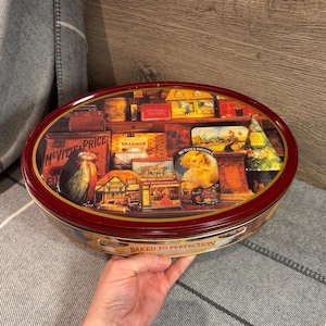 Puede incluir: Una lata vintage roja y dorada con una ilustración colorida de una tienda de panadería y el texto "Baked to Perfection".