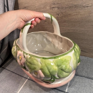 Puede incluir: Una cesta de cerámica con asa, pintada con flores rosas y hojas verdes. La cesta tiene un asa blanca y una base rosa. La cesta está sujeta por una mano.