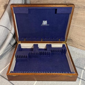 以下が含まれることがあります： 「Prince Silver」とラベルが貼られた、青いベルベットの内装と木製のシルバーウェアチェスト。チェストには、シルバーウェアを収納するための複数のコンパートメントがあります。