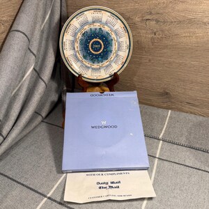 May include: A Wedgwood plate with a calendar design for 2005, in blue and white. The plate is displayed on a wooden stand, with a blue Wedgwood box and a Daily Mail document visible below.