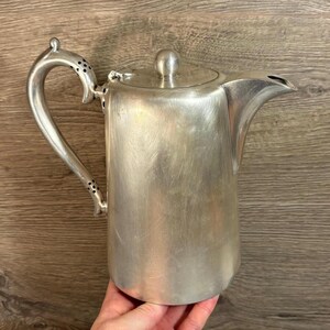 Puede incluir: Una jarra de plata con un pico redondeado y un asa con agujeros decorativos. La jarra tiene una superficie lisa y ligeramente texturizada.