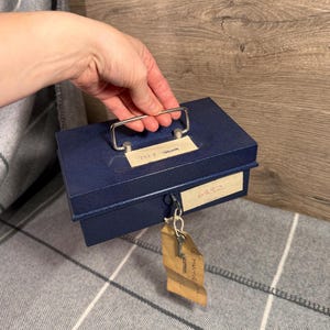 Może przedstawiać: Granatowa metalowa kasa z srebrnym uchwytem i kluczem wiszącym na zamku. Pudełko ma dwie białe etykiety z numerami i tekstem. Pudełko znajduje się na szaro-białym pledzie w kratę.