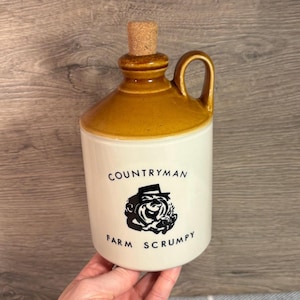Könnte beinhalten: Eine weiße Keramikkanne mit braunem Deckel und einem Korkstopfen. Die Kanne hat einen Henkel und zeigt eine Schwarzweißillustration eines Mannes mit Hut und dem Text "COUNTRYMAN FARM SCRUMPY".