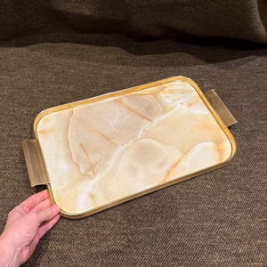 Puede incluir: Una bandeja de servicio rectangular con una superficie de piedra veteada en tonos crema y bronceado. La bandeja está enmarcada por un borde de metal dorado pulido e incluye dos asas de metal dorado a juego con una textura estriada vertical.