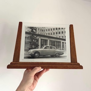 Könnte beinhalten: Schwarz-Weiß-Fotografie eines Oldtimers in einem Holzrahmen. Der Rahmen hat eine dunkelbraune Oberfläche und wird gehalten. Das Foto zeigt einen Oldtimer, der vor einem Gebäude geparkt ist.