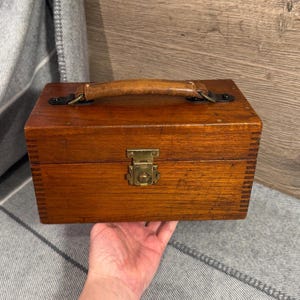 May include: A rectangular wooden box with a leather handle and brass fittings. The box has a hinged lid and a clasp. The wood is a warm, reddish-brown colour, and the box shows signs of wear, indicating it is an antique or vintage item.