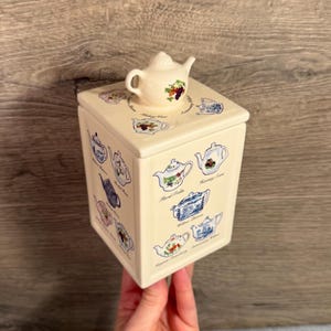 May include: A cream-colored square ceramic canister featuring a lid topped with a miniature teapot handle. The canister is decorated with various colorful teapot illustrations and labels including Floral Trellis, Hunting Scene, and Willow Pattern.