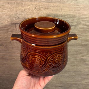 May include: A brown ceramic jar with a lid and two handles. The jar has a swirling pattern on the body.