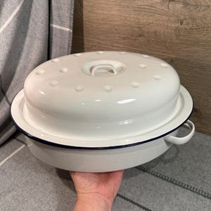 Könnte beinhalten: Weißer Emaille-Bräter mit Deckel. Die Pfanne hat einen dunkelblauen Rand und Griffe. Der Deckel hat einen Griff und kleine, erhabene Punkte. Die Pfanne steht auf einer grau-weiß gestreiften Decke.