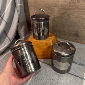 May include: Three dark grey metal canisters with lids and handles. One reads "Sugar", another "Tea", and the third "Coffee". The canisters have a hammered texture and a vintage aesthetic.