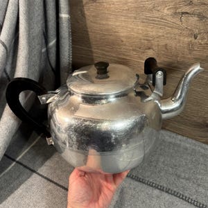 Op de afbeelding: Een zilveren aluminium theepot met een zwarte handgreep en tuit. De theepot heeft een glanzende afwerking en een licht gedeukte uitstraling.