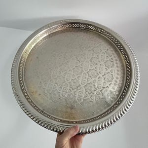 May include: A round, silver-coloured metal tray with an intricate, embossed geometric pattern. The tray has a decorative, scalloped edge and a raised rim. The tray is held by a hand.