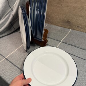 Pode incluir: Um conjunto de pratos redondos brancos com borda azul-escura. V&aacute;rios pratos est&atilde;o empilhados verticalmente num suporte de madeira, enquanto um prato individual &eacute; segurado em primeiro plano, exibindo o acabamento liso e a borda azul-escura.