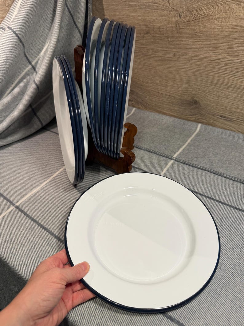 Pode incluir: Um conjunto de pratos redondos brancos com borda azul-escura. V&aacute;rios pratos est&atilde;o empilhados verticalmente num suporte de madeira, enquanto um prato individual &eacute; segurado em primeiro plano, exibindo o acabamento liso e a borda azul-escura.