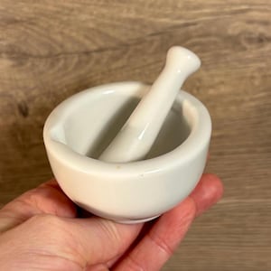 Könnte beinhalten: Ein weißer Keramikmörser und Stößel. Der Mörser ist rund und hat eine glatte, weiße Oberfläche. Der Stößel ist zylindrisch und passt perfekt in den Mörser.