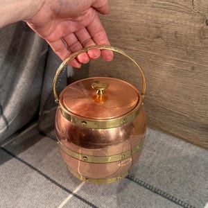 Op de afbeelding: Een koperen ijsemmer met een deksel en een messing handvat. De emmer heeft messing banden rond het midden en is geklonken. De handgreep is gebogen en getextureerd. De emmer is rond en heeft een glanzende afwerking.