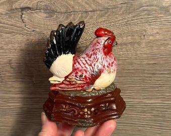 Peut inclure: Figurine de coq en céramique avec une crête et des barbillons rouges, un corps moucheté de rouge et de blanc, et des plumes de queue noires et dorées. Le coq est posé sur un socle décoratif marron avec des accents dorés.