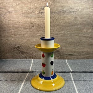 Op de afbeelding: Een gele keramische kaarsenstandaard met een witte basis versierd met kleurrijke fruitontwerpen. De standaard heeft een blauwe rand en een blauwe band rond de basis. Een witte kaars staat in de standaard.