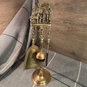 Puede incluir: Un juego de herramientas para chimenea de latón con un soporte decorativo que presenta un relieve de una casa y figuras. El juego incluye una pala, un cepillo y un atizador.