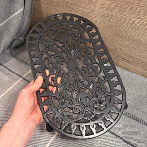 May include: Dark grey cast iron trivet with an oval shape and an elaborate tree and leaf pattern. The trivet has small feet and a decorative border. The trivet measures approximately 25 cm long and 15 cm wide.