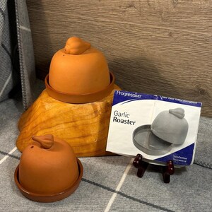 May include: Two terracotta garlic roasters with lids shaped like garlic cloves. The roasters are on a wooden base. The packaging for the roasters is in the background. The packaging is blue and white and says "Progressive Garlic Roaster".