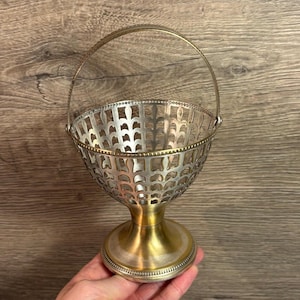 May include: A silver and gold metal basket with a decorative cut-out design and a handle. The basket is on a pedestal base.