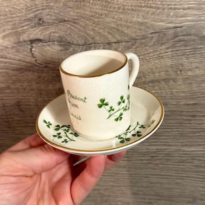 May include: A small, white teacup and saucer set with gold trim. The cup has a handle and the words "Present from Ennis" in green script. Green shamrock designs decorate the cup and saucer.