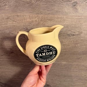 Op de afbeelding: Een crèmekleurige keramische kan met een zwart ovaal label met witte tekst die luidt: Fine Single Malt, Aged 10 Years, Tamdhu, Product of Scotland, Scotch Whisky. De kan heeft een gebogen handvat en een schenktuit aan de rand.