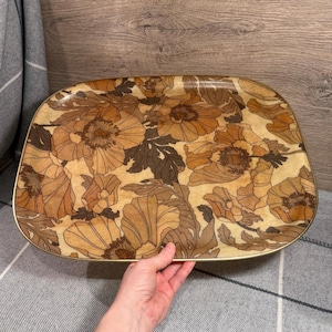 Op de afbeelding: Rechthoekig dienblad met een bloemenpatroon in bruin-, beige- en goudtinten. De lade heeft een goudkleurige rand en een vintage uitstraling. Het bloemmotief toont grote, gestileerde bloemen en bladeren.