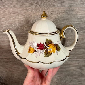 Puede incluir: Una tetera blanca con ribete dorado y diseño floral con rosas rojas y amarillas. La tetera tiene una tapa con un pomo dorado.