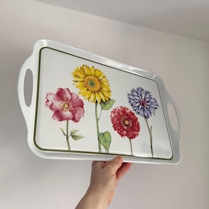 Può includere: Vassoio rettangolare bianco con un motivo floreale. Il vassoio presenta un girasole, una rosa rosa, un papavero rosso e un fiordaliso blu. Il vassoio ha un bordo verde e maniglie integrate.
